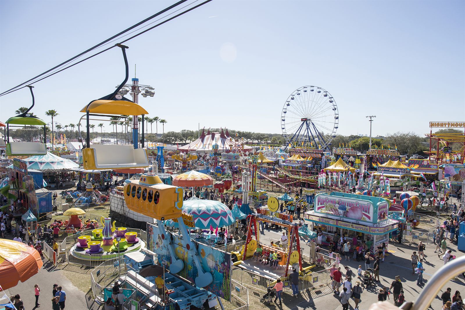 A busy fair midway seen from above, with a sky ride, colorful rides and a Ferris wheel
